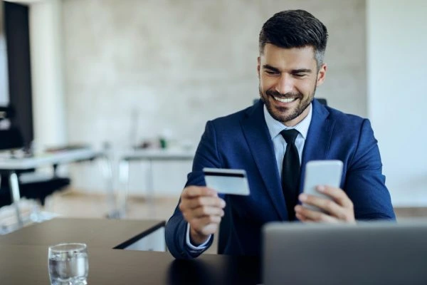 A business professional seated at a desk uses a smartphone while holding a payment card, with a laptop open in front and a glass of water nearby.