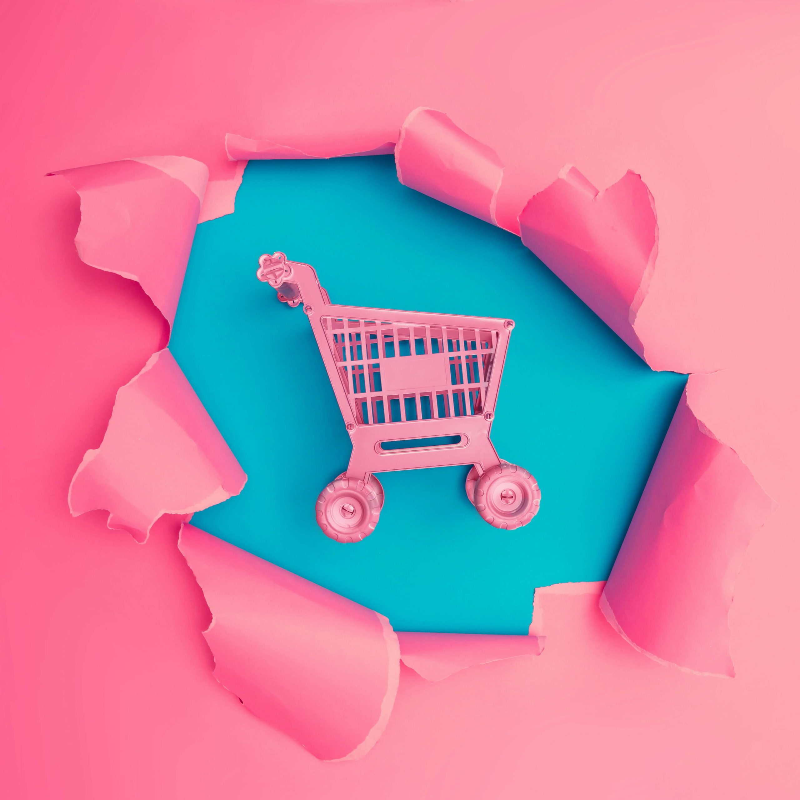 Pink plastic toy shopping cart tearing through bright pink paper, revealing a teal blue background behind it.