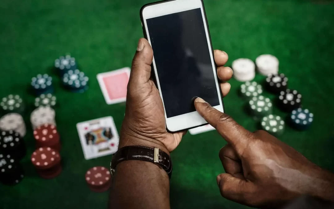 Man using a smartphone for online gaming with poker chips and playing cards on a green casino table.