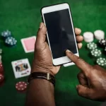Man using a smartphone for online gaming with poker chips and playing cards on a green casino table.