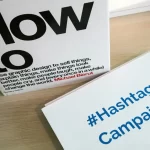 Book How to by Michael Bierut alongside a card reading “#Hashtag Campaign” on a wooden surface.